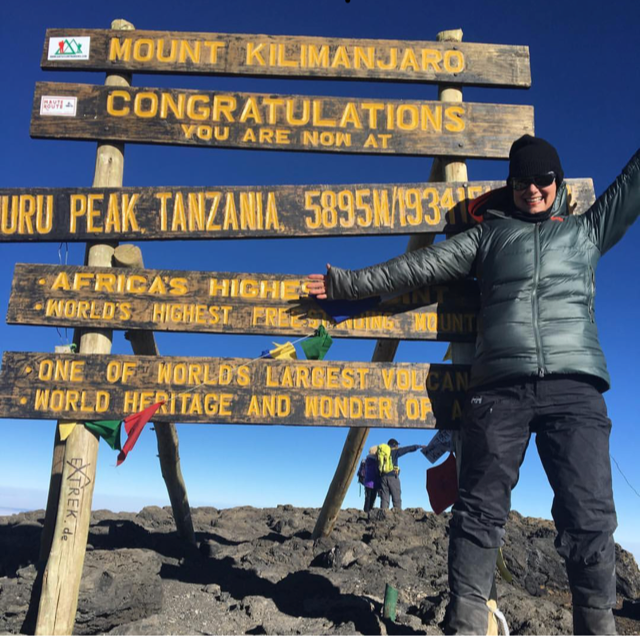 A woman stands in a black puffy and beanie in front of the Kilimanjaro summit sign.