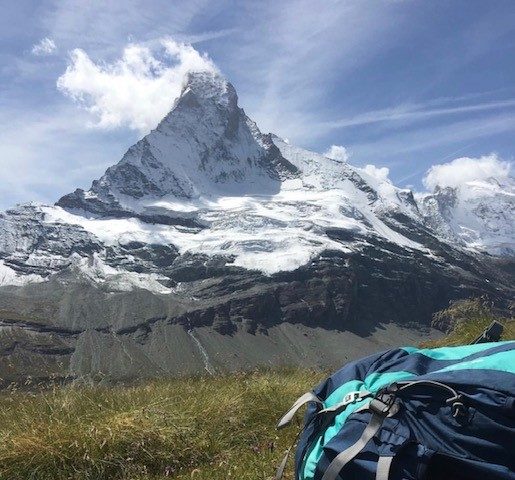 The Matterhorn rises in the background. The midground is a verdant meadow. The foreground is a backpack.