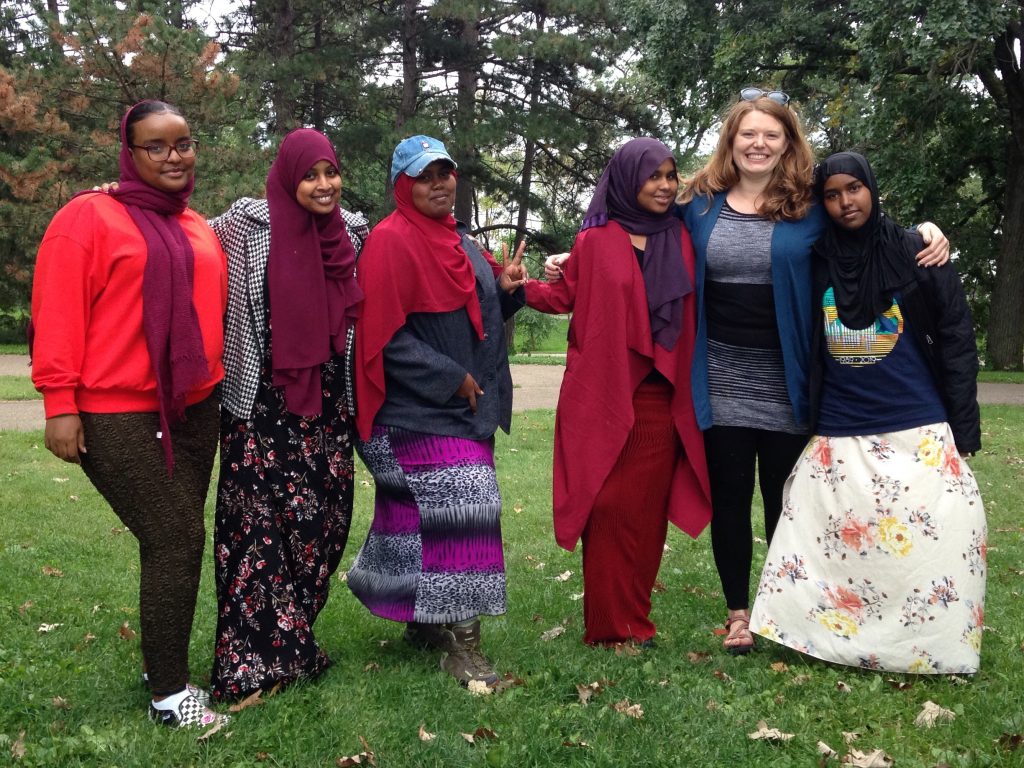 Five students stand smiling in a park. Their Trip Instructor is embracing two of them.