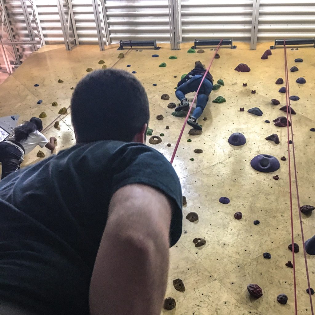 The student in the foreground is belaying the student in the background, who is nearly to the top of an indoor rock wall.