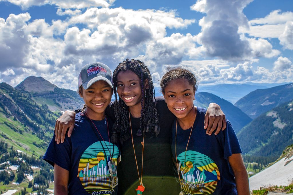 Three young women stand in front of a mountainous backdrop. They have their arms around each other, and are smiling for the camera.