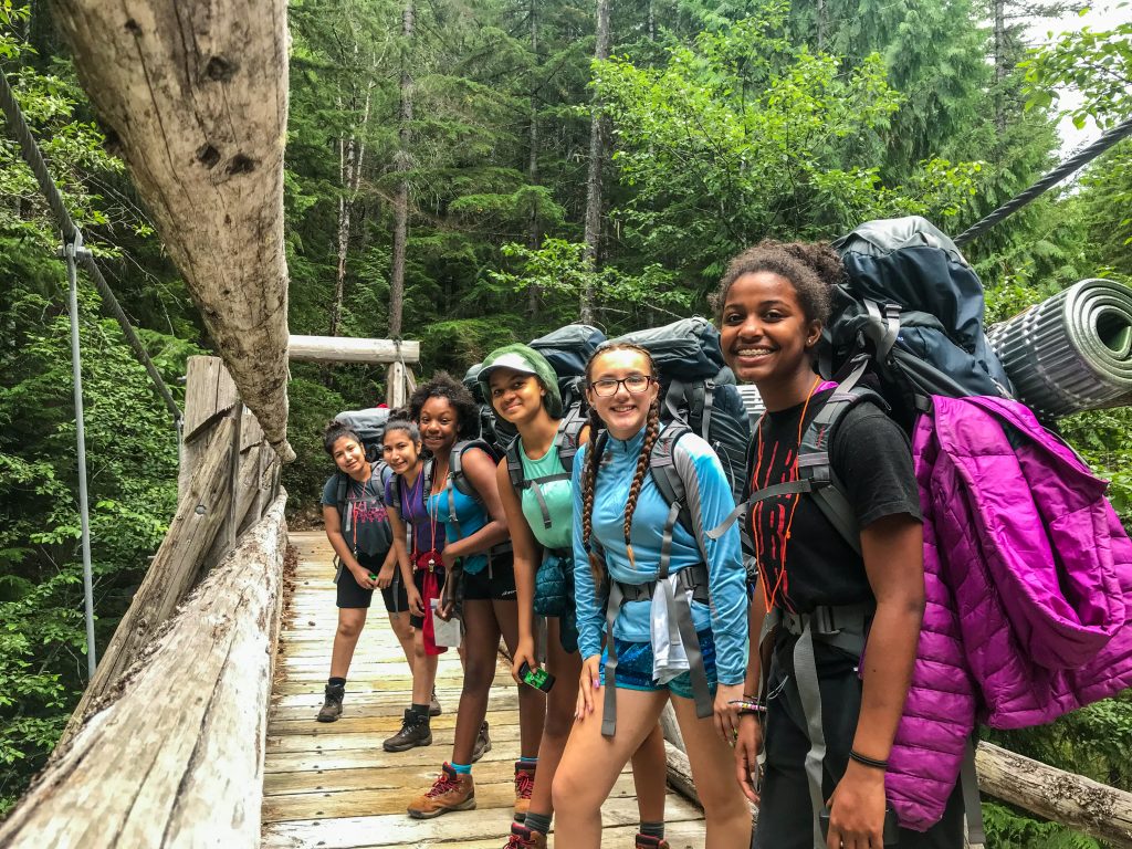 Six youth with backpacking gear stand in an angled line on a wooden bridge. A dense forest stands in the background.